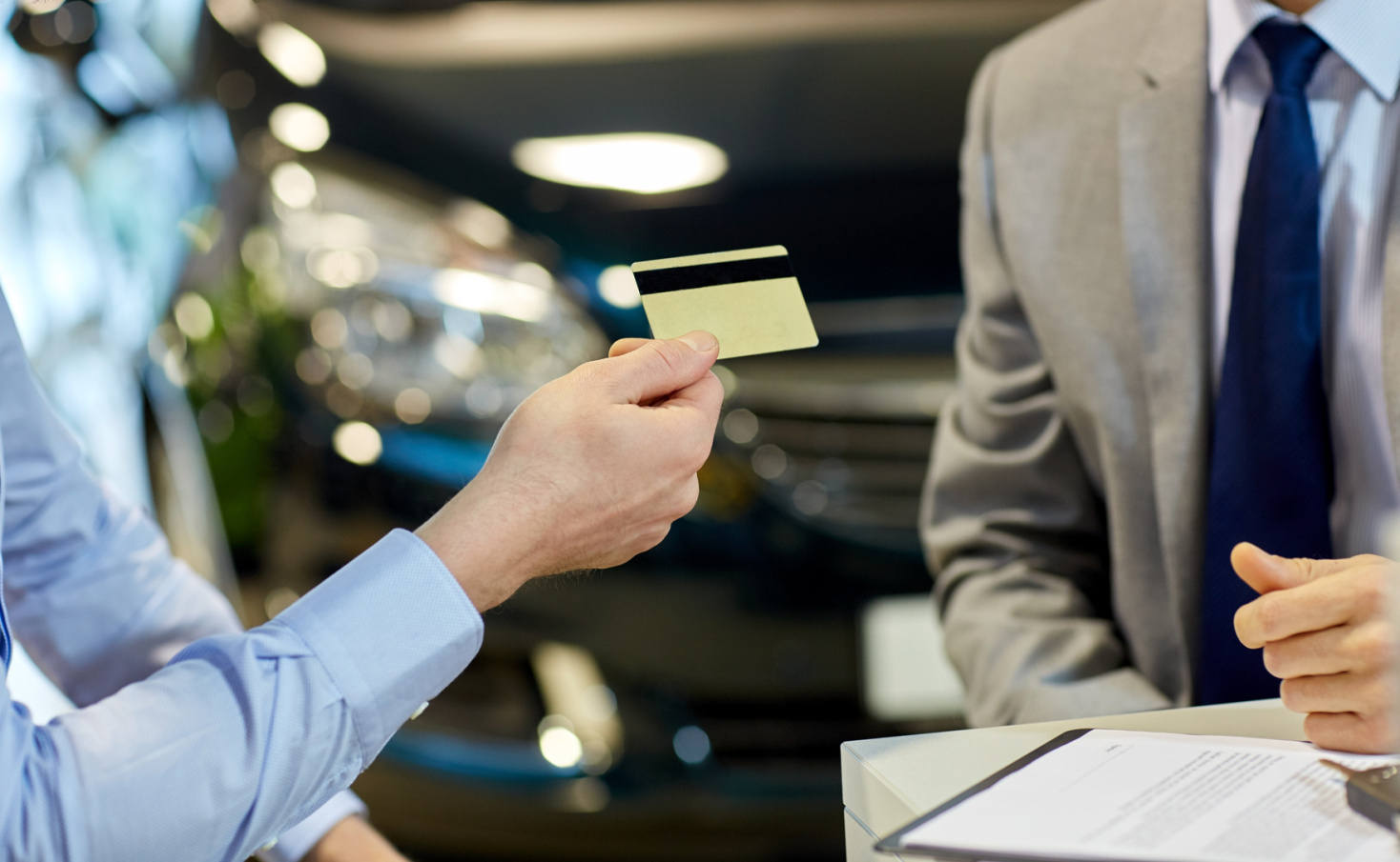 Zwei Männer sitzen über einem Vertrag in einem Autohaus, der Interessent hält dem Verkäufer eine Kreditkarte hin; Kurtz Wirtschaftsdetektei Nürnberg.